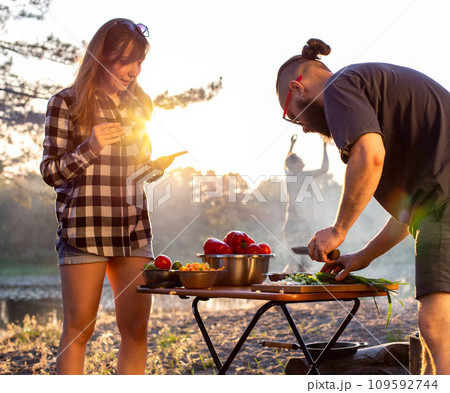 A brutal caucasian man with a beard and black glasses is preparing a vegetable salad in nature against the backdrop of an evening sunset. The girl shoots on the phone. Cooking in nature, tourism A brutal caucasian man with a beard and black glasses is preparing a vegetable salad in nature against the backdrop of an evening sunset. The girl shoots on the phone. Cooking in nature, tourism 109592744