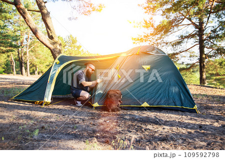 A handsome guy with a beard and black glasses sets up a tourist tent on the sandy bank of the river in the forest. Outdoor recreation in summer. Hiking, hobby, travel. Copy space for text A handsome guy with a beard and black glasses sets up a tourist tent on the sandy bank of the river in the forest. Outdoor recreation in summer. Hiking, hobby, travel. Copy space for text 109592798