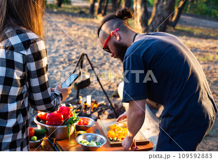 Stylish man with a beard and black glasses cuts fresh vegetables peppers and tomatoes to prepare a salad in nature. Vacation with friends in nature. Tourism as a hobby. Evening sunset. Stylish man with a beard and black glasses cuts fresh vegetables peppers and tomatoes to prepare a salad in nature. Vacation with friends in nature. Tourism as a hobby. Evening sunset. 109592883