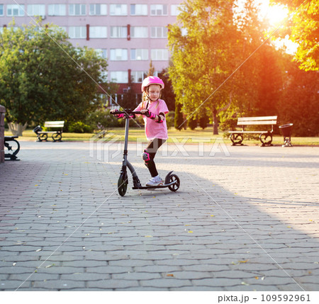 A girl 7 years old in a helmet and a pink injury protection kit rides a scooter in the park, sunny day. Active sports and recreation. Safety when riding bicycles and scooters. Copy space for text A girl 7 years old in a helmet and a pink injury protection kit rides a scooter in the park, sunny day. Active sports and recreation. Safety when riding bicycles and scooters. Copy space for text 109592961