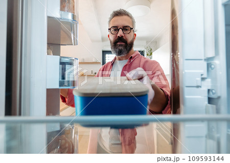 Handsome man putting lunchbox in fridge. Taking out lunch from fridge, eating leftovers. Stop trowing food away, reducting food waste. 109593144