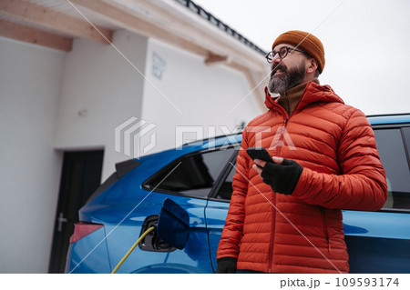 Man charging electric car during cold snowy day, using electric vehicle charging app, checking battery life, energy consumption on smart phone. Charging and driving electric vehicles during winter 109593174
