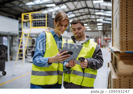 Young man with Down syndrome working in warehouse, female colleague teaching him scan products with scanner. Concept of workers with disabilities, support in workplace. 109593247