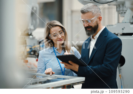Female engineer and male production manager standing in modern industrial factory, talking about production. Manufacturing facility with robotics and automation. 109593290