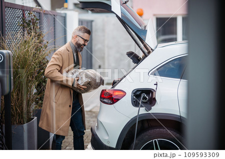 Man putting bag of wood pellets in car trunk, while his electric vehicle is charging. 109593309