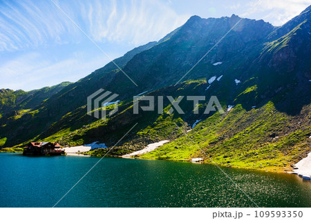 mountainous summer landscape of romania. green scenery of balea lake in carpathian fagaras ridge on a sunny morning. fluffy clouds on the bright blue sky. spots of snow on the grassy hillsides mountainous summer landscape of romania. green scenery of balea lake in carpathian fagaras ridge on a sunny morning. fluffy clouds on the bright blue sky. spots of snow on the grassy hillsides 109593350
