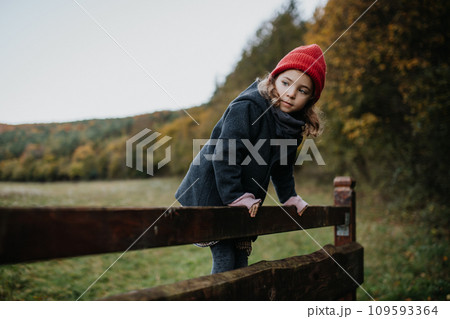 Young girl in warm clothes sitting on aged wooden fence, wooden gate, looking at autumn nature. 109593364