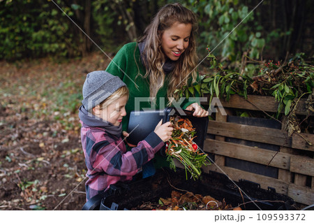 Boy helping mother to put kitchen waste, peel and leftover vegetables scraps into composter in the garden. Concept of composting kitchen biodegradable waste. 109593372