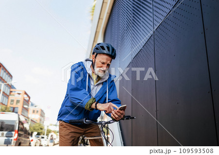 Elderly man, cyclist traveling through the city by bike, reading text message on smartphone. Senior city commuter riding a bike to grocery store, checking shopping list in mobile app. 109593508