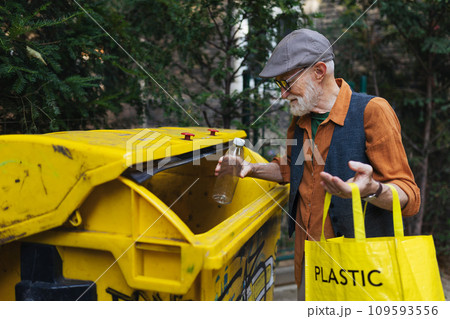 Senior man throwing plastic waste, bottles into recycling container in front apartment. Elderly man sorting the waste according to material into colored bins. Senior man throwing plastic waste, bottles into recycling container in front apartment. Elderly man sorting the waste according to material into colored bins. 109593556