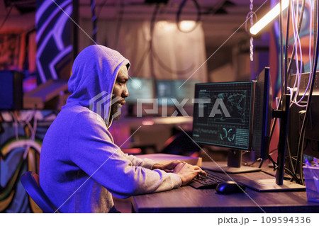 African american hacker coding spyware in abandoned warehouse. Man in hood using computer for breaching data, cracking password and getting access to server database at night 109594336