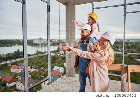 Man holding daughter and woman pointing at something while standing next to husband at construction site. Family with child future homeowners observing apartment building under construction. Man holding daughter and woman pointing at something while standing next to husband at construction site. Family with child future homeowners observing apartment building under construction. 109597453