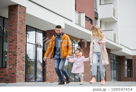 Full length of man and woman holding hands of little girl while walking down the street in new urban district. Happy loving family with child enjoying stroll outdoors near residential building. 109597454