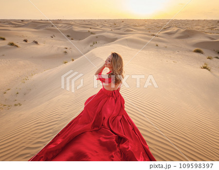 Desert adventure. Young arabian Woman in red silk dress in sands dunes of UAE desert at sunset, fantastic view. The Dubai Desert Conservation Reserve, United Arab Emirates. Desert adventure. Young arabian Woman in red silk dress in sands dunes of UAE desert at sunset, fantastic view. The Dubai Desert Conservation Reserve, United Arab Emirates. 109598397