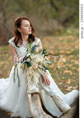 beautiful bride wearing white boots sitting on tree and holding autumn bouquet. 109600003