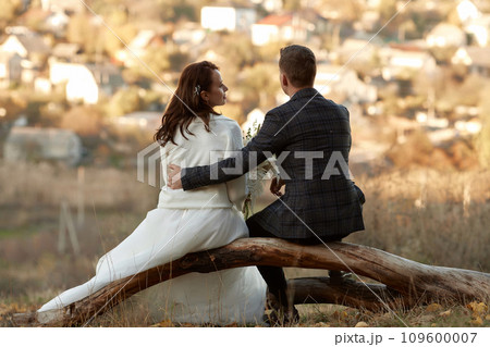 bride in white wedding dress and groom are sitting on tree in nature, back view 109600007