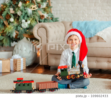 Happy little boy with a gift, toy train, under the Christmas tree on New Year's morning. Time to fulfill wishes. Happy little boy with a gift, toy train, under the Christmas tree on New Year's morning. Time to fulfill wishes. 109602433