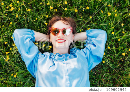 Top view happy young woman in blue blouse lays on a green grass lawn with yellow flowers, looks up and smiles. Business woman enjoy the moment, relaxing outdoors. Spring or summer season concept. 109602943