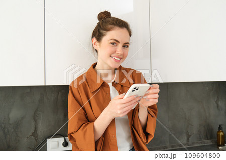 Portrait of young smiling woman with mobile phone, standing in the kitchen, looking happy and relaxed at camera Portrait of young smiling woman with mobile phone, standing in the kitchen, looking happy and relaxed at camera 109604800