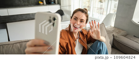 Friendly smiling young woman, sitting on couch and waving hand at smartphone camera, video chats, talking on mobile phone, connects to online conversation on application 109604824