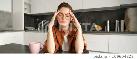 Portrait of working woman at home, student has headache, touches head and has painful migraine, sits in kitchen in glasses 109604825