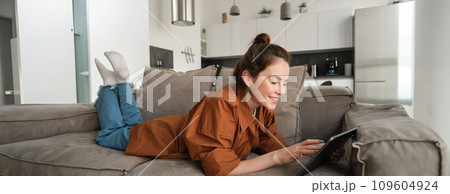 Portrait of beautiful young woman resting at home on couch, lying on sofa with digital tablet, reading or watching videos on gadget Portrait of beautiful young woman resting at home on couch, lying on sofa with digital tablet, reading or watching videos on gadget 109604924