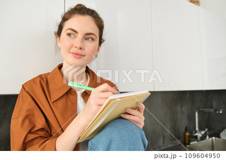 Portrait of woman at home, sitting with notebook and pen, writing, making notes, looking outside with thoughtful smiling face 109604950