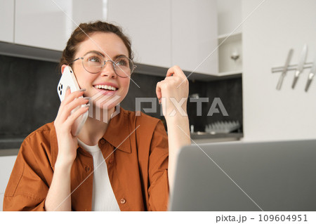 Portrait of young woman at home, sitting in kitchen with laptop, talking on mobile phone, making a call, working in her flat 109604951