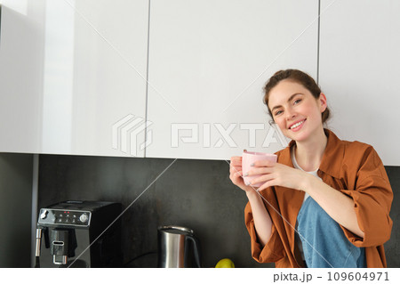 Portrait of beautiful young woman enjoying her morning cup of aromatic coffee, drinking tea while sitting in kitchen and contemplating sunny day 109604971