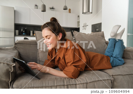 Portrait of beautiful young woman resting at home on couch, lying on sofa with digital tablet, reading or watching videos on gadget Portrait of beautiful young woman resting at home on couch, lying on sofa with digital tablet, reading or watching videos on gadget 109604994
