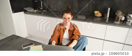 Portrait of beautiful young woman studying at home, sitting with laptop and documents in kitchen. Self-employed business owner typing on computer, sending emails to clients Portrait of beautiful young woman studying at home, sitting with laptop and documents in kitchen. Self-employed business owner typing on computer, sending emails to clients 109605010
