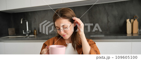 Close up portrait of cute and tender young woman in glasses, sitting in kitchen, drinking tea, holding cup, tucks her hair behind ear and smiles 109605100