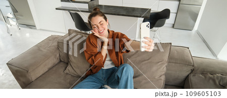 Portrait of beautiful smiling woman, takes selfie at home, posing on sofa, holds smartphone with extended hand 109605103