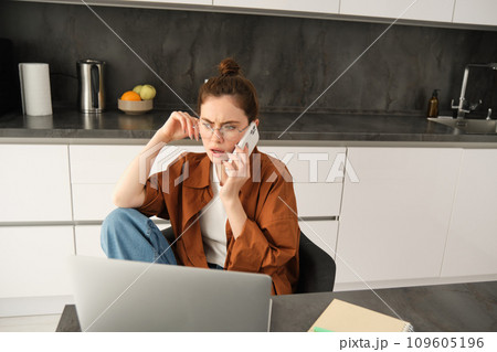 Portrait of busy young self-employed woman, working from home, has office in her flat, sitting near laptop, answers phone call, talking with serious, concerned face expression Portrait of busy young self-employed woman, working from home, has office in her flat, sitting near laptop, answers phone call, talking with serious, concerned face expression 109605196