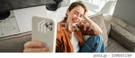 Carefree modern woman, smiling and taking selfies on smartphone app, posing for photo on her couch, sitting on sofa with mobile phone 109605199