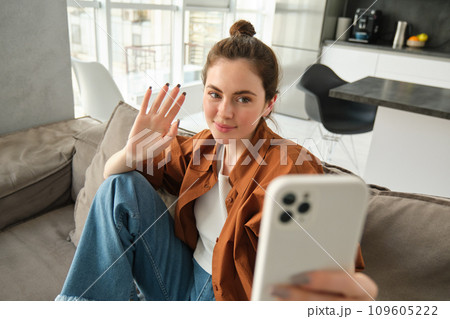 Happy woman is sitting at home with smartphone, online chatting, waving hand at mobile phone, saying hello, talking to friends using application, sitting on sofa at home 109605222