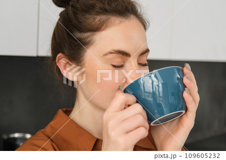 Portrait of beautiful young woman, enjoying delicious aroma of freshly made coffee, holding mug, standing near machine in the kitchen, drinking from mug Portrait of beautiful young woman, enjoying delicious aroma of freshly made coffee, holding mug, standing near machine in the kitchen, drinking from mug 109605232