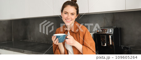 Portrait of beautiful young woman, enjoying delicious aroma of freshly made coffee, holding mug, standing near machine in the kitchen, drinking from mug Portrait of beautiful young woman, enjoying delicious aroma of freshly made coffee, holding mug, standing near machine in the kitchen, drinking from mug 109605264