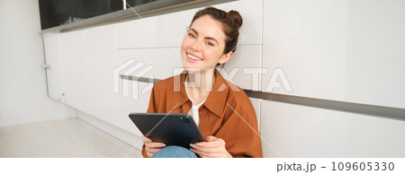 Young entrepreneurs. Smiling cute woman with digital tablet, sitting with gadget on kitchen floor and looking happy, studying remotely, reading on her device 109605330