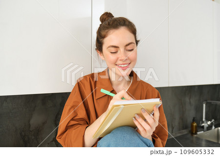 Portrait of young creative girl, writing down her ideas in planner, does homework in notebook, sits at home on kitchen counter Portrait of young creative girl, writing down her ideas in planner, does homework in notebook, sits at home on kitchen counter 109605332