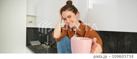 Pretty young smiling woman sits in kitchen, offers you cup of coffee, extends hand with pink mug, sharing her drink with someone 109605738
