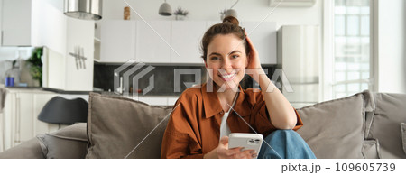 Beautiful smiling female model sitting on couch with smartphone, looking happy at camera, scrolling social media, messaging someone, doing online shopping 109605739
