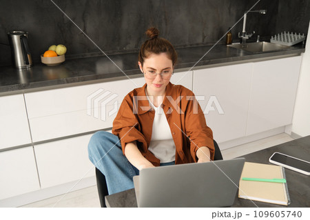 Portrait of beautiful young woman studying at home, sitting with laptop and documents in kitchen. Self-employed business owner typing on computer, sending emails to clients Portrait of beautiful young woman studying at home, sitting with laptop and documents in kitchen. Self-employed business owner typing on computer, sending emails to clients 109605740
