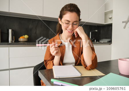 Young woman making notes, sitting in kitchen with pen and notebook, smiling while writing in her planner, planning her schedule 109605761