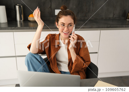 Excited young woman talking on mobile phone in front of laptop, sitting in kitchen with happy face expression, having a conversation Excited young woman talking on mobile phone in front of laptop, sitting in kitchen with happy face expression, having a conversation 109605762