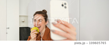 Portrait of happy, smiling young woman records herself, takes selfie while eating an apple in the kitchen, using smartphone app, makes photos with mobile phone 109605765