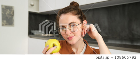 Beautiful young woman in glasses, sitting on chair in the kitchen, eating green apple and smiling Beautiful young woman in glasses, sitting on chair in the kitchen, eating green apple and smiling 109605792