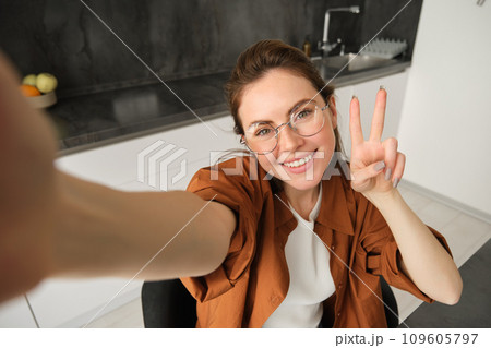 Portrait of stylish young woman at home, taking selfie on smartphone, posing in kitchen and smiling at mobile camera 109605797