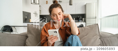 Young woman with calm, relaxed face, reading on smartphone, looking at mobile phone screen, sitting on couch in living room Young woman with calm, relaxed face, reading on smartphone, looking at mobile phone screen, sitting on couch in living room 109605893