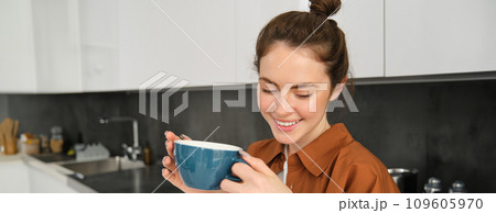 Portrait of beautiful young woman, enjoying delicious aroma of freshly made coffee, holding mug, standing near machine in the kitchen, drinking from mug 109605970
